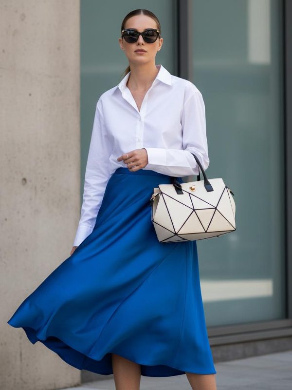 Ein asymmetrischer Rock in Kobaltblau verbindet sich mit einem weissen Oversize-Hemd, einer geometrischen Tasche und einer Sonnenbrille. Ein starker Farbton, der durch den weiten Schnitt seine Strenge verliert.